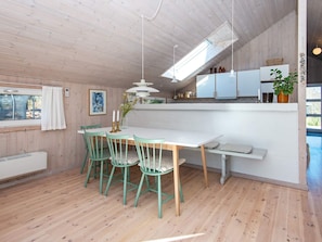A dining area with a white table and green chairs, a kitchen with cabinets and a hanging light, and a window with a curtain.