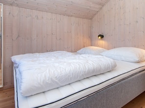 Un lit simple avec un matelas et des oreillers blancs, dans une pièce aux murs en bois et dotée d’un luminaire au plafond.