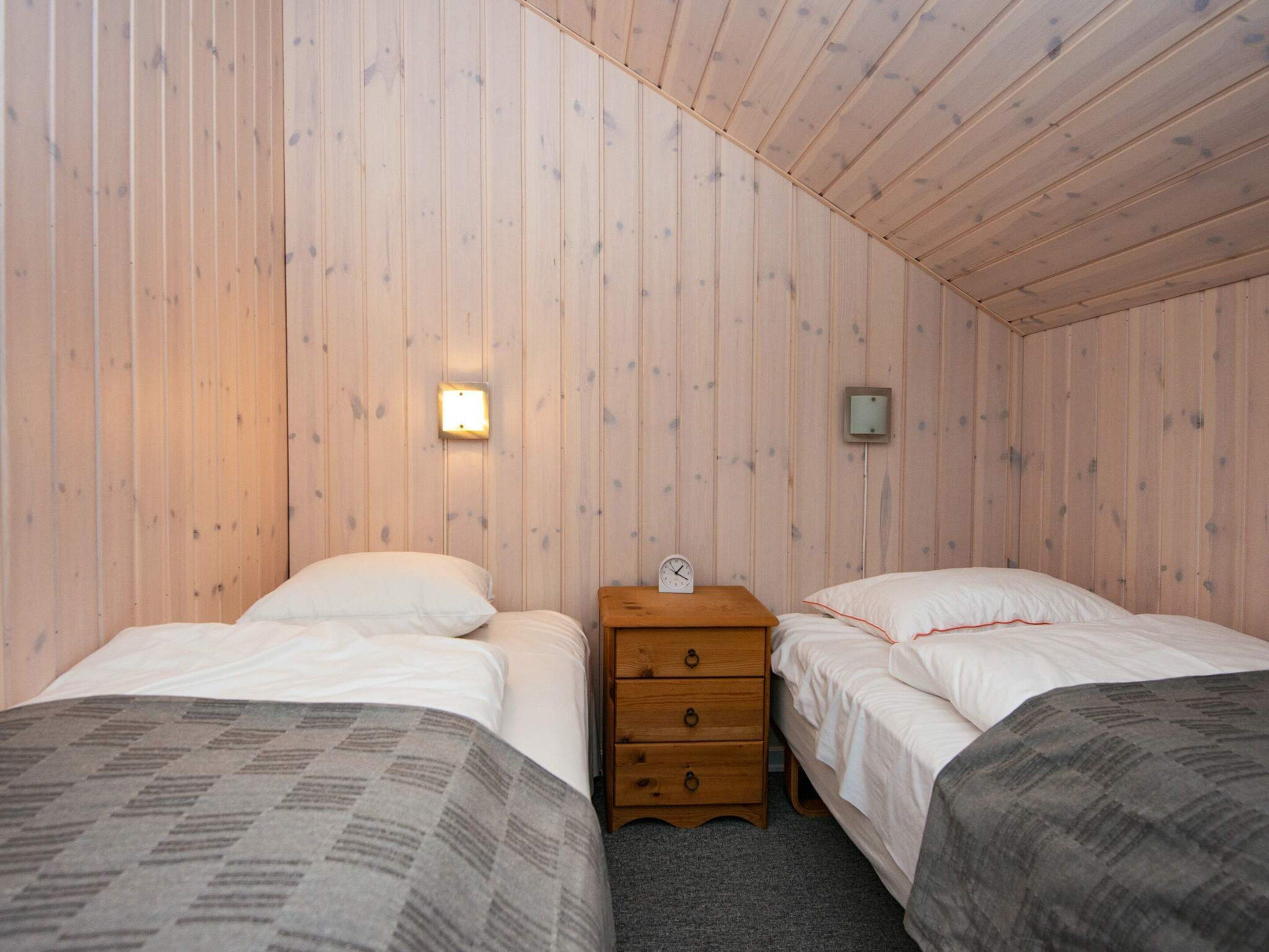 A room with two beds, a wooden nightstand with a clock, and a wall-mounted light fixture.