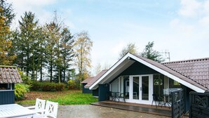 A covered patio area with a table and chairs, a small building with a tiled roof, and a view of trees and a grassy area. - 7 Person Holiday Home in Ansager (Ansager)