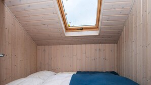 Quarto com teto inclinado, paredes de madeira, claraboia e cama com roupa de cama branca e azul.