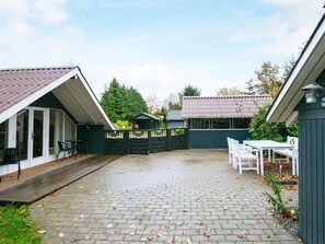 Área de varanda coberta com piso de tijolos, deck de madeira e jardim cercado.