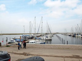 Un puerto deportivo con varias embarcaciones amarradas, una orilla pedregosa y algunas personas caminando por el muelle.
