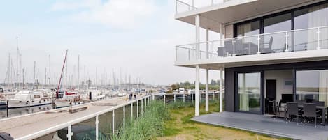 A modern building with a glass facade, a balcony with outdoor seating, and a view of a marina with boats.