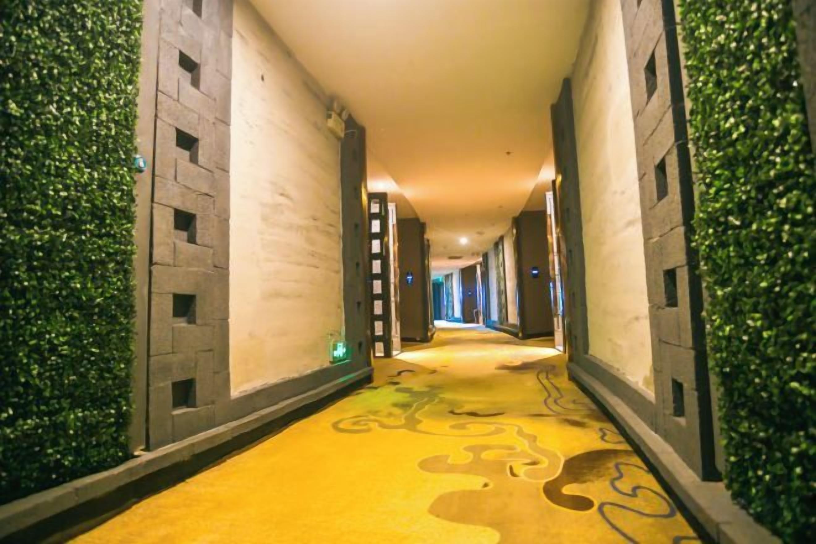 A hotel corridor with a patterned carpet, walls with vertical lines, and green foliage on the sides.