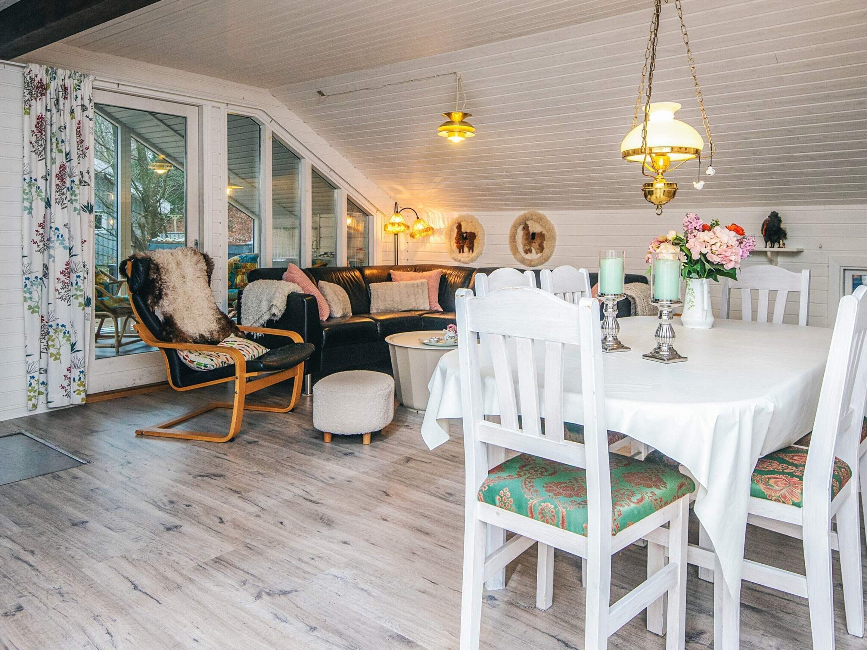 A cozy dining area with a white table set for four, surrounded by white chairs with patterned cushions. A black leather sofa with a fluffy throw and a wooden rocking chair are positioned against the wall. The room features a wooden ceiling with hanging pendant lights, and a large window with a view of the outdoors.