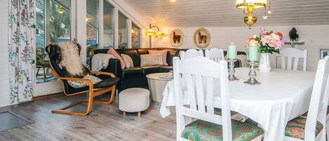 A cozy dining area with a white table set for four, surrounded by white chairs with patterned cushions. A black leather sofa with a fluffy throw and a wooden rocking chair are positioned against the wall. The room features a wooden ceiling with hanging pendant lights, and a large window with a view of the outdoors.