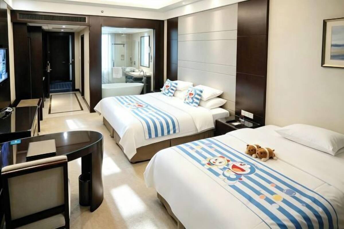 Habitación de hotel con dos camas, un escritorio, una silla y un baño visible a través de una puerta abierta.