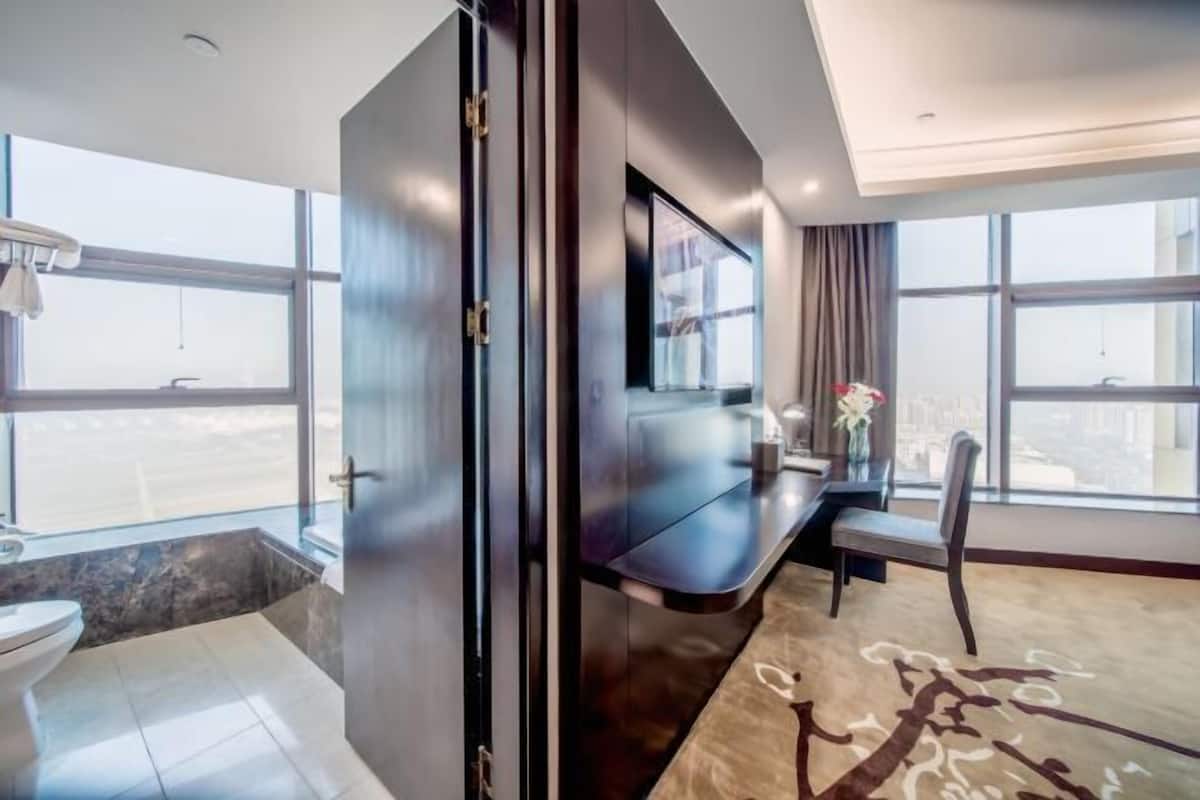 A modern hotel room with a large window, a flat-screen TV, a desk, and a chair.