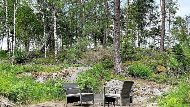En utomhusplats med sittmöbler, bestående av två fåtöljer i rotting och en bänk, omgiven av en skogbeklädd sluttning.