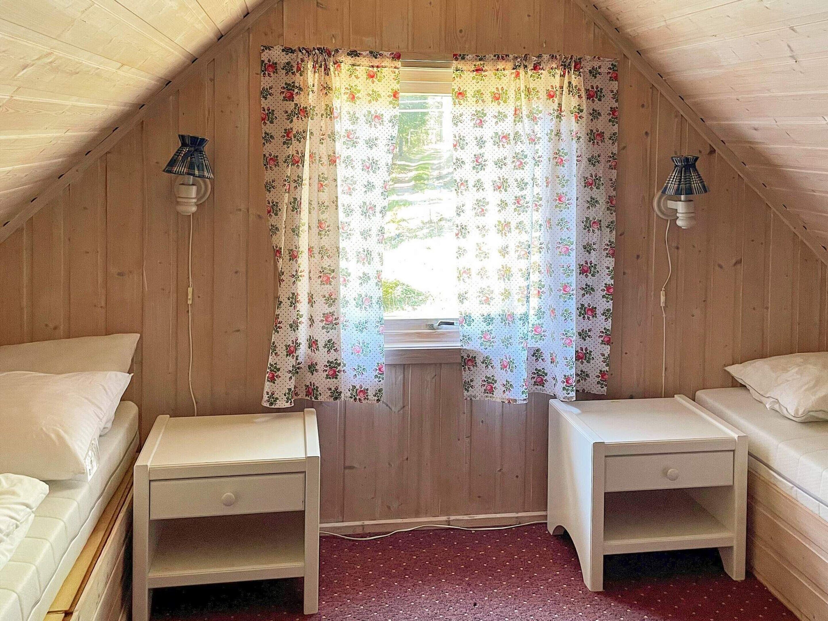 A small, cozy attic bedroom with two single beds, wooden walls, a floral curtain, and two wall-mounted lamps.