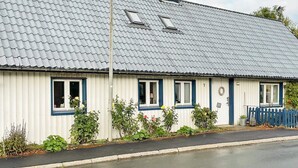 Un edificio de una sola planta con techo de metal corrugado, paredes exteriores blancas y detalles azules alrededor de las ventanas. Hay un pequeño jardín con diversas plantas y flores frente al edificio.