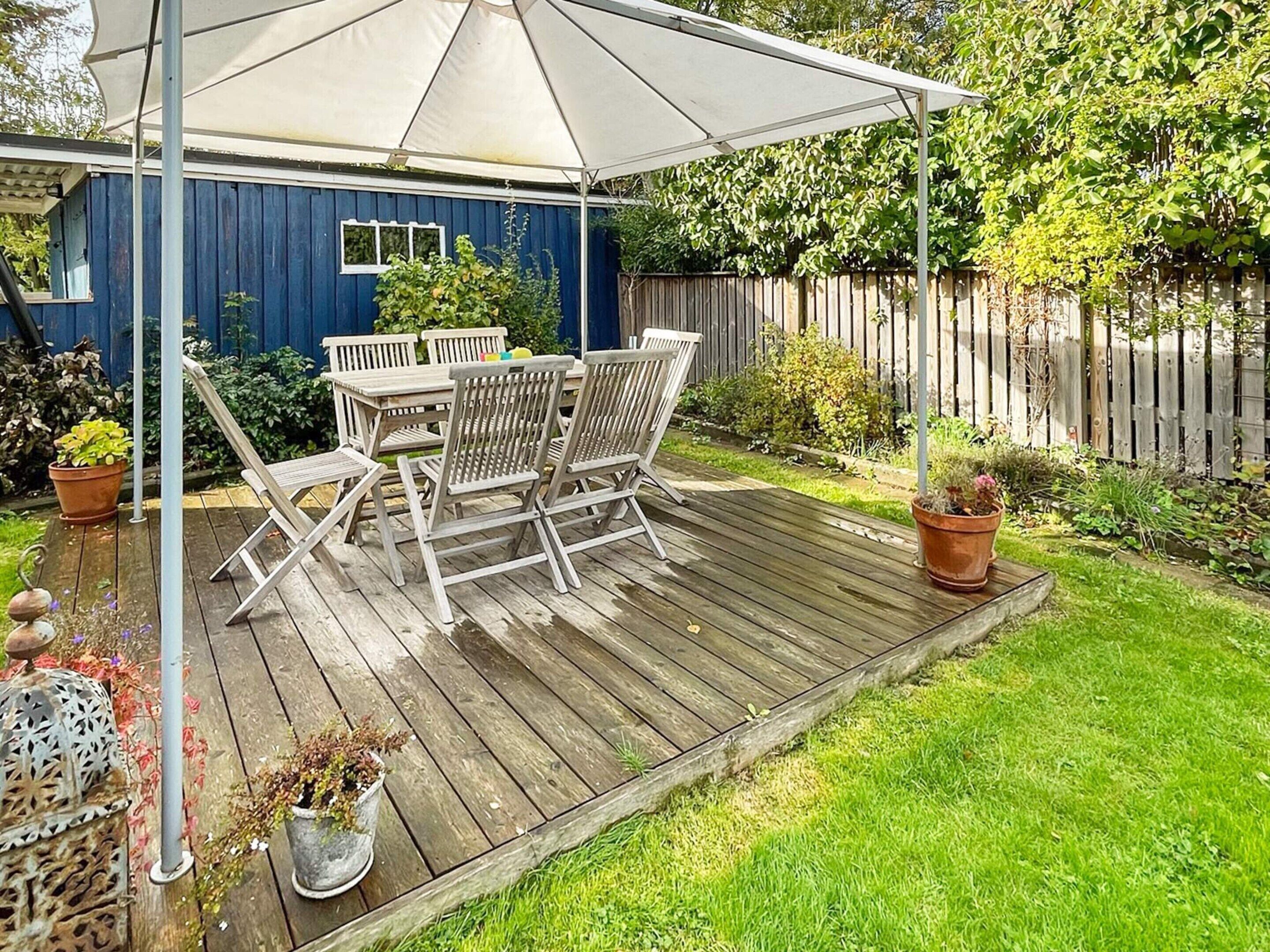 Una terraza de madera con una mesa y sillas bajo un paraguas blanco, rodeada de plantas en macetas y una valla de madera.