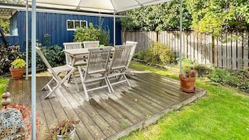 Una terraza de madera con una mesa y sillas bajo un paraguas blanco, rodeada de plantas en macetas y una valla de madera.