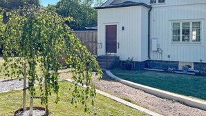 Une maison blanche à deux étages, avec une porte d’entrée, un petit porche et un jardin avec un arbre.