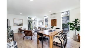 Uma sala ampla com uma mesa de jantar de madeira, cadeiras pretas e um sofá. Há grandes janelas que oferecem luz natural e vista para o exterior.