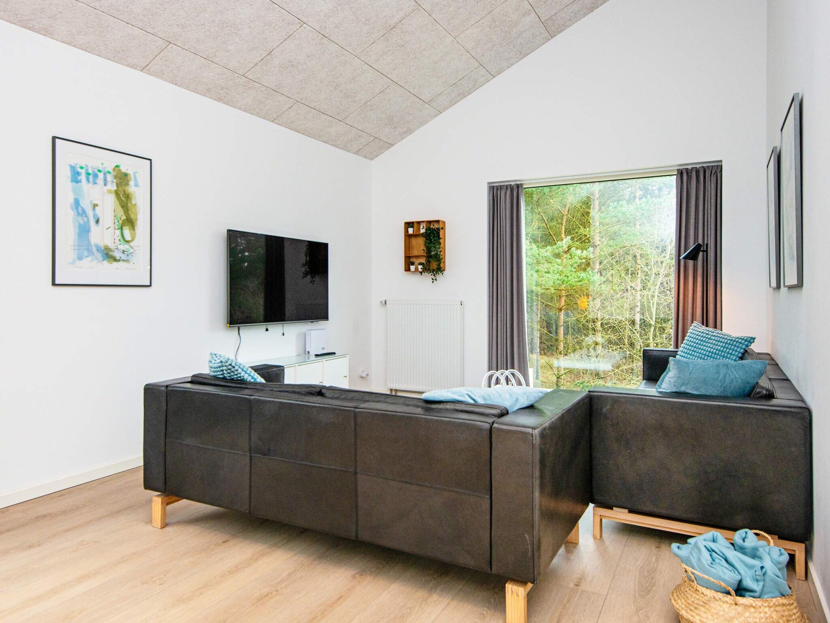 A modern living room with a sectional sofa, a flat-screen TV, a large window with curtains, and a framed artwork on the wall.