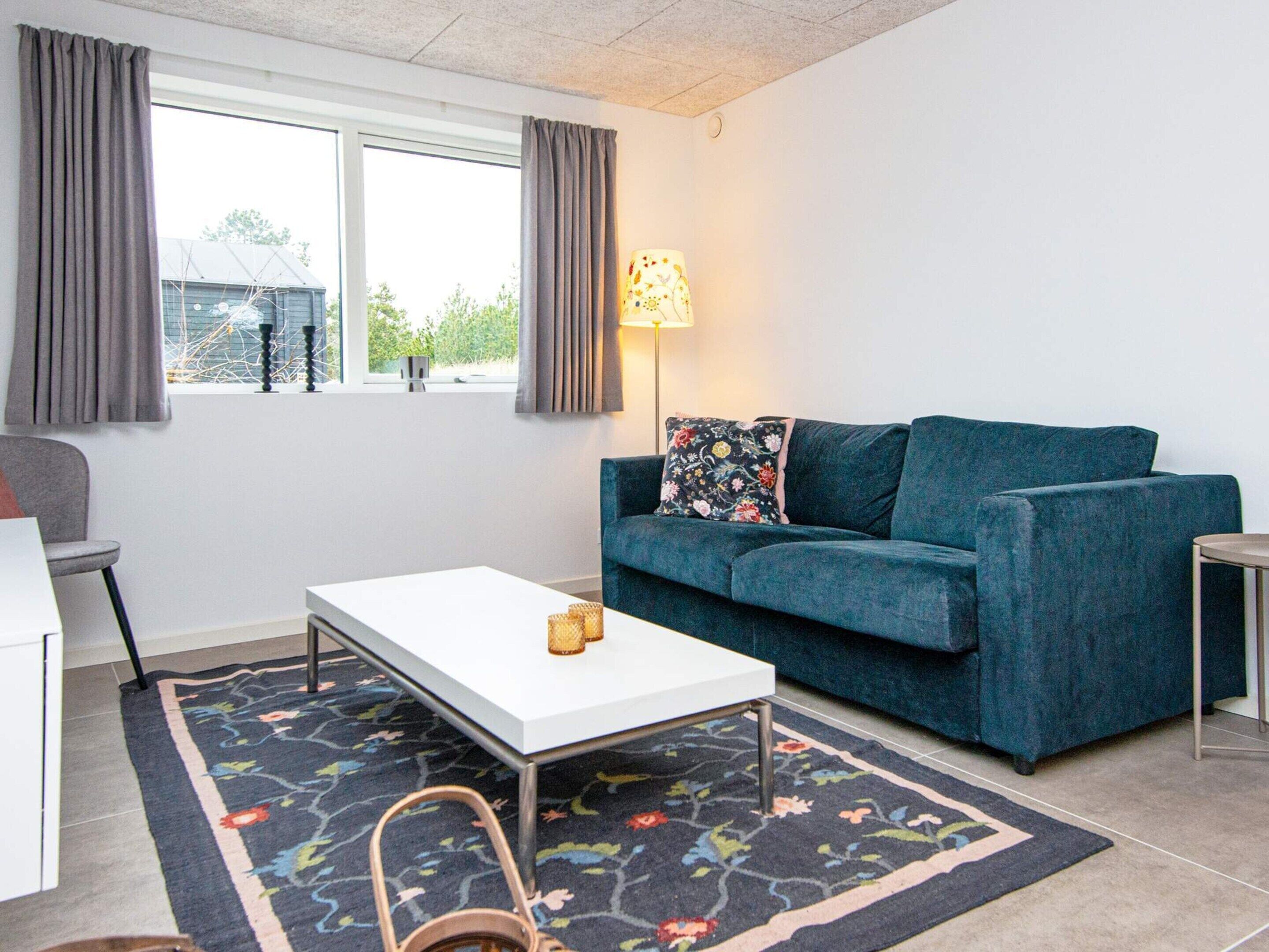 Ein Wohnzimmer mit einem blauen Sofa, einem weißen Couchtisch, einem gemusterten Teppich und einem Fenster mit Vorhängen.