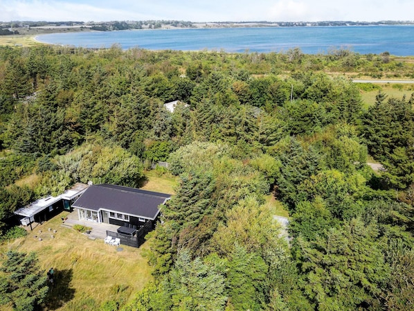 A single-story house surrounded by dense forest, with a view of a large body of water in the background.