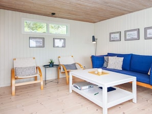 A room with a wooden ceiling, white walls, a blue sofa, two wooden chairs, a white coffee table, and framed pictures on the wall.