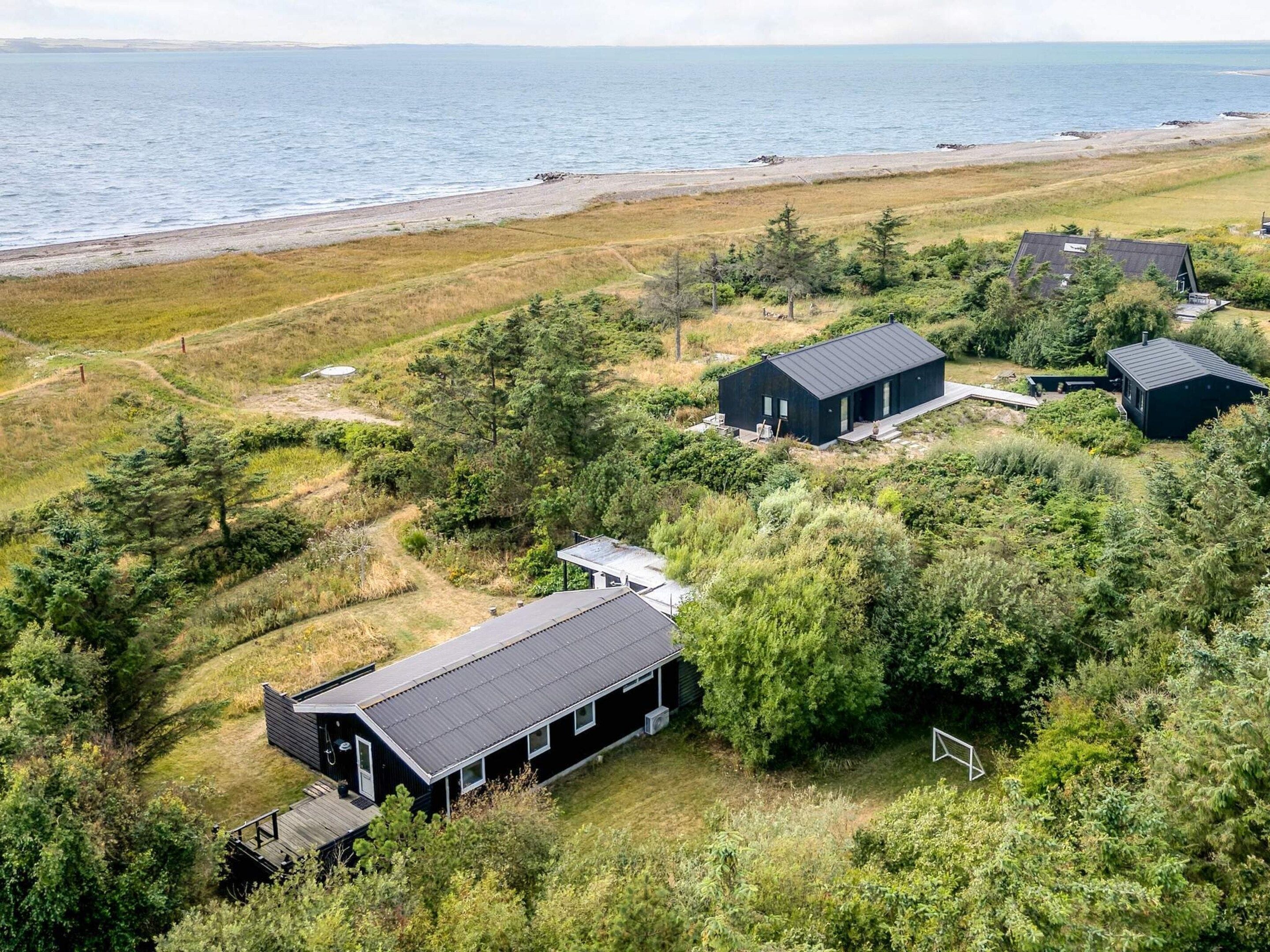 Una propiedad costera con varias construcciones, incluyendo una casa grande con porche cubierto y una estructura más pequeña, rodeada de vegetación densa y una playa al fondo.
