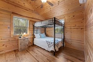 A wooden cabin bedroom with a bunk bed, a bedside table with a lamp, and a window overlooking a forest.