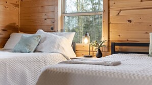 A cozy bedroom with two beds, a bedside table with a lamp, and a window with a view of trees.