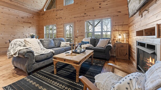 A cozy log cabin interior with a fireplace, two sofas, a coffee table, and large windows.