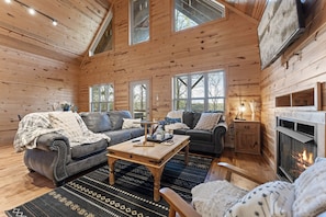 A cozy log cabin interior with a fireplace, two sofas, a coffee table, and large windows.