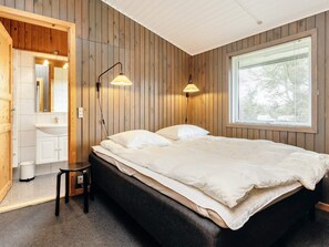 A bedroom with a wooden wall, a bed, a bedside table, a chair, a window, and a bathroom with a mirror and sink. - 8 Person Holiday Home in Lokken (Lokken)