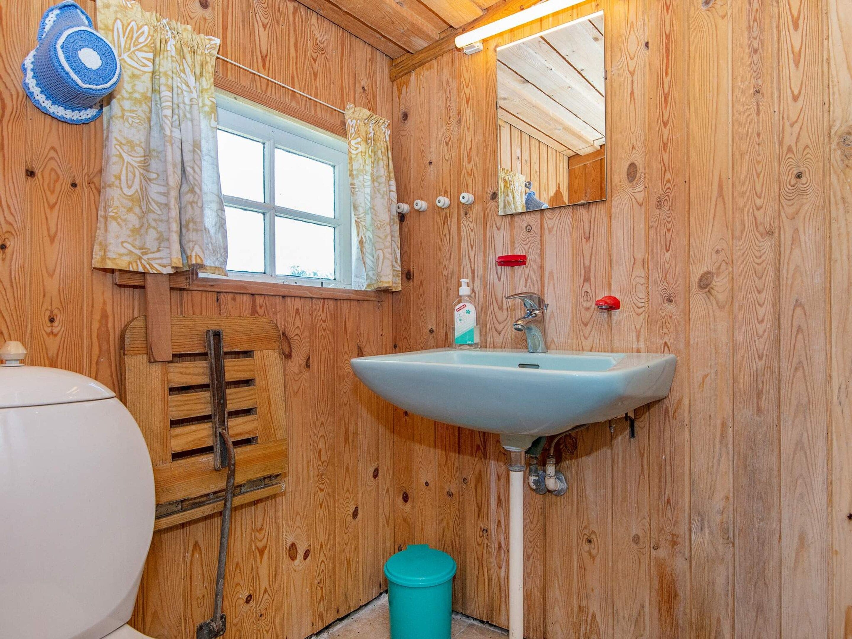 Une salle de bain en bois avec un lavabo blanc, un miroir, une fenêtre avec des rideaux et des toilettes.