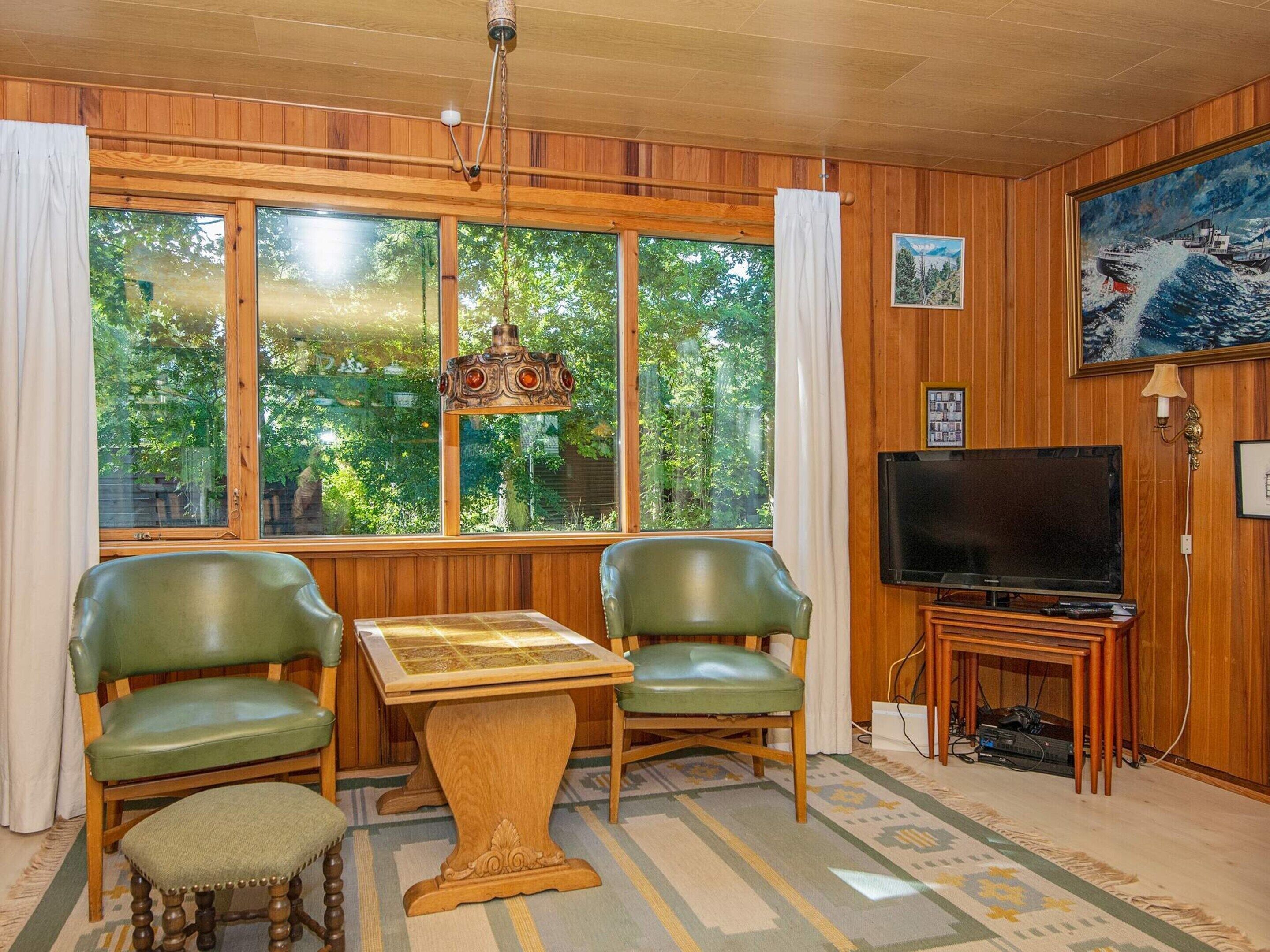 Una habitación con paneles de madera, televisor, dos sillas verdes, una mesita y un gran ventanal con cortinas.