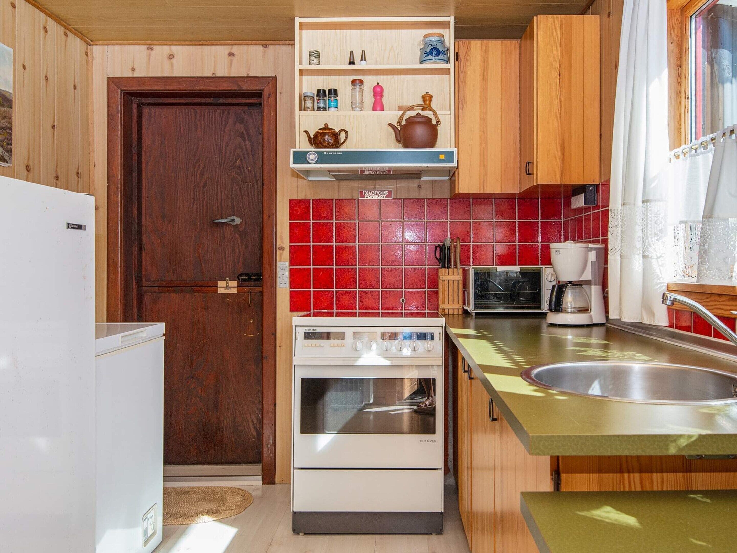 A compact kitchen with a wooden door, a white refrigerator, a built-in oven, a coffee maker, and a red-tiled backsplash.