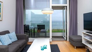Ein modernes Wohnzimmer mit einer Couch, einem Couchtisch und einem Fernseher. Eine Glastür öffnet sich zu einem Balkon mit Blick auf den Ozean.