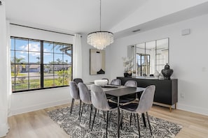Une salle à manger moderne avec une grande fenêtre, un lustre, une table à manger avec des chaises et une console sombre agrémentée d’objets décoratifs.