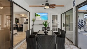 A dining area with a glass partition, wicker furniture, a ceiling fan, and a TV displaying a beach scene. - Canal View! Walking distance to Marina Village Fully Renovated (Cape Coral)