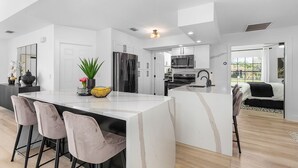 A modern kitchen with white cabinetry, a central island with bar stools, and a large window providing natural light. - Canal View! Walking distance to Marina Village Fully Renovated (Cape Coral)