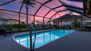 An outdoor swimming pool with a glass enclosure, surrounded by lounge chairs and palm trees, under a sunset sky. - Canal View! Walking distance to Marina Village Fully Renovated (Cape Coral)