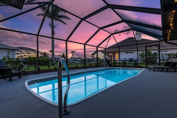 Une piscine extérieure dotée d’une structure en verre, entourée de chaises longues et de palmiers, sous un ciel au coucher du soleil.