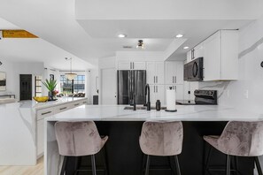 Une cuisine moderne avec des meubles blancs, un grand îlot central doté de tabourets, des appareils électroménagers en acier inoxydable et une vue sur l’extérieur.