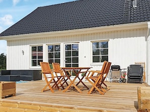 A white house with a dark roof, a deck with wooden chairs and a table, and a barbecue grill.