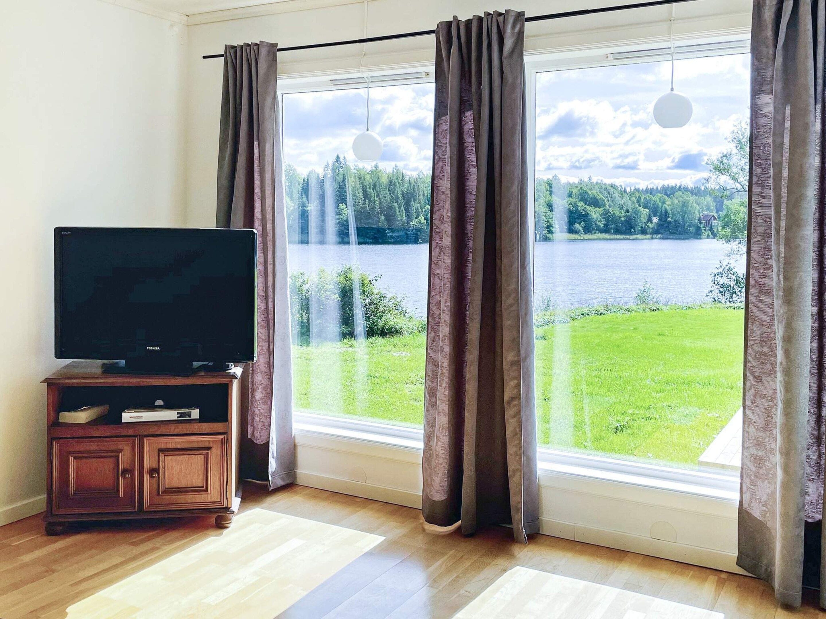 A room with a television on a wooden cabinet, large windows with curtains, and a view of a lake and forest.