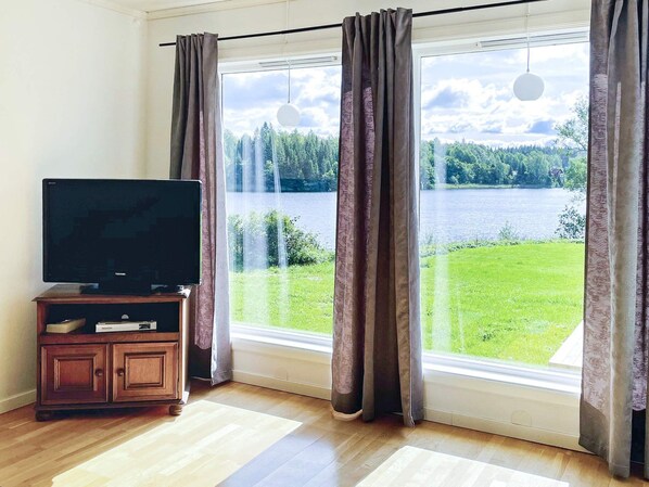Una habitación con una televisión sobre un mueble de madera, ventanas amplias con cortinas y vistas a un lago y un bosque.