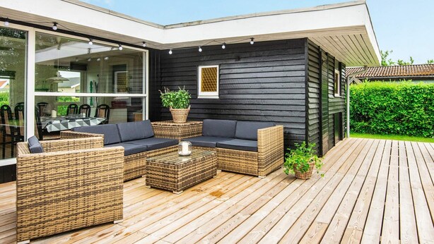 Ett utomhusområde för avkoppling och umgänge med ett modernt hus, som inkluderar en träveranda, möbler i rotting och en glasdörr som leder till en matplats.