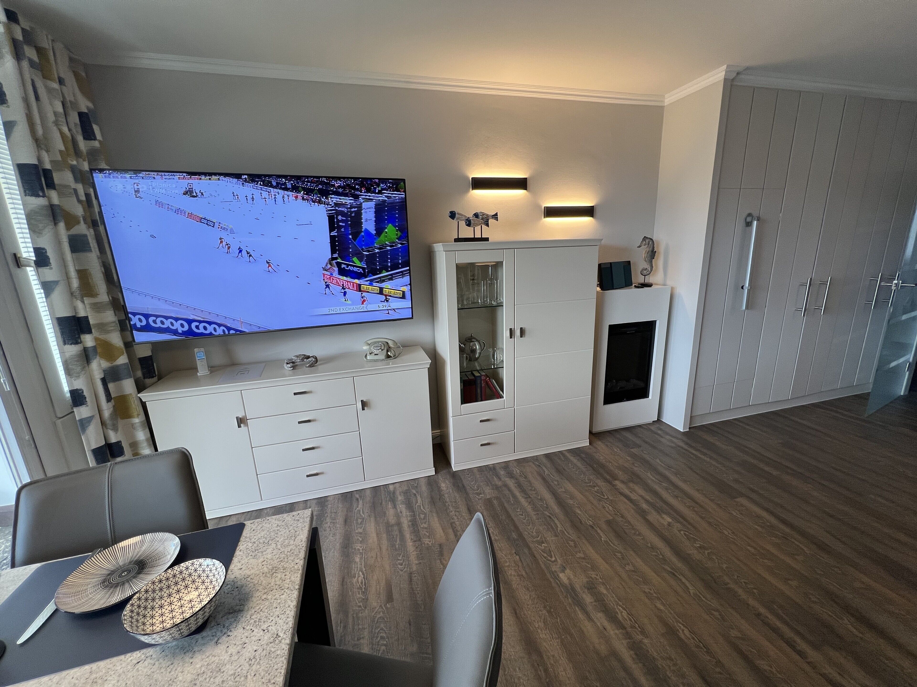A modern kitchen with a flat-screen TV, white cabinetry, and a dining area with a table and chairs.