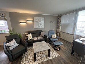 Un salon moderne comprenant un canapé, deux fauteuils, une table basse et une table d’appoint. On y trouve une grande fenêtre avec des stores, un radiateur et une œuvre d’art accrochée au mur.