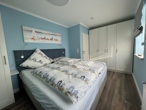 Une chambre à coucher comprenant un lit, une tête de lit, une œuvre d’art accrochée au mur, une armoire et une fenêtre avec des stores.