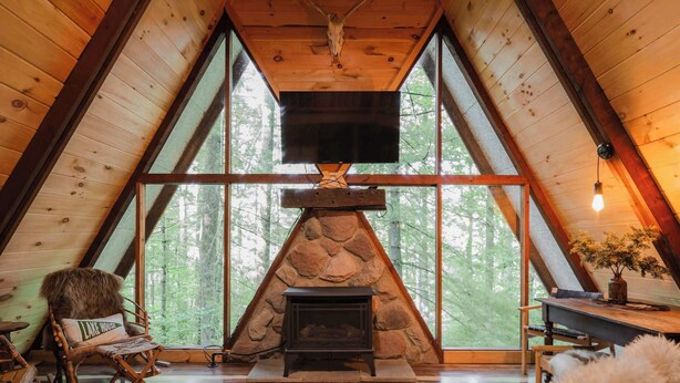 L’intérieur chaleureux d’un chalet avec une cheminée en pierre, des murs en bois et de grandes fenêtres donnant sur la forêt.