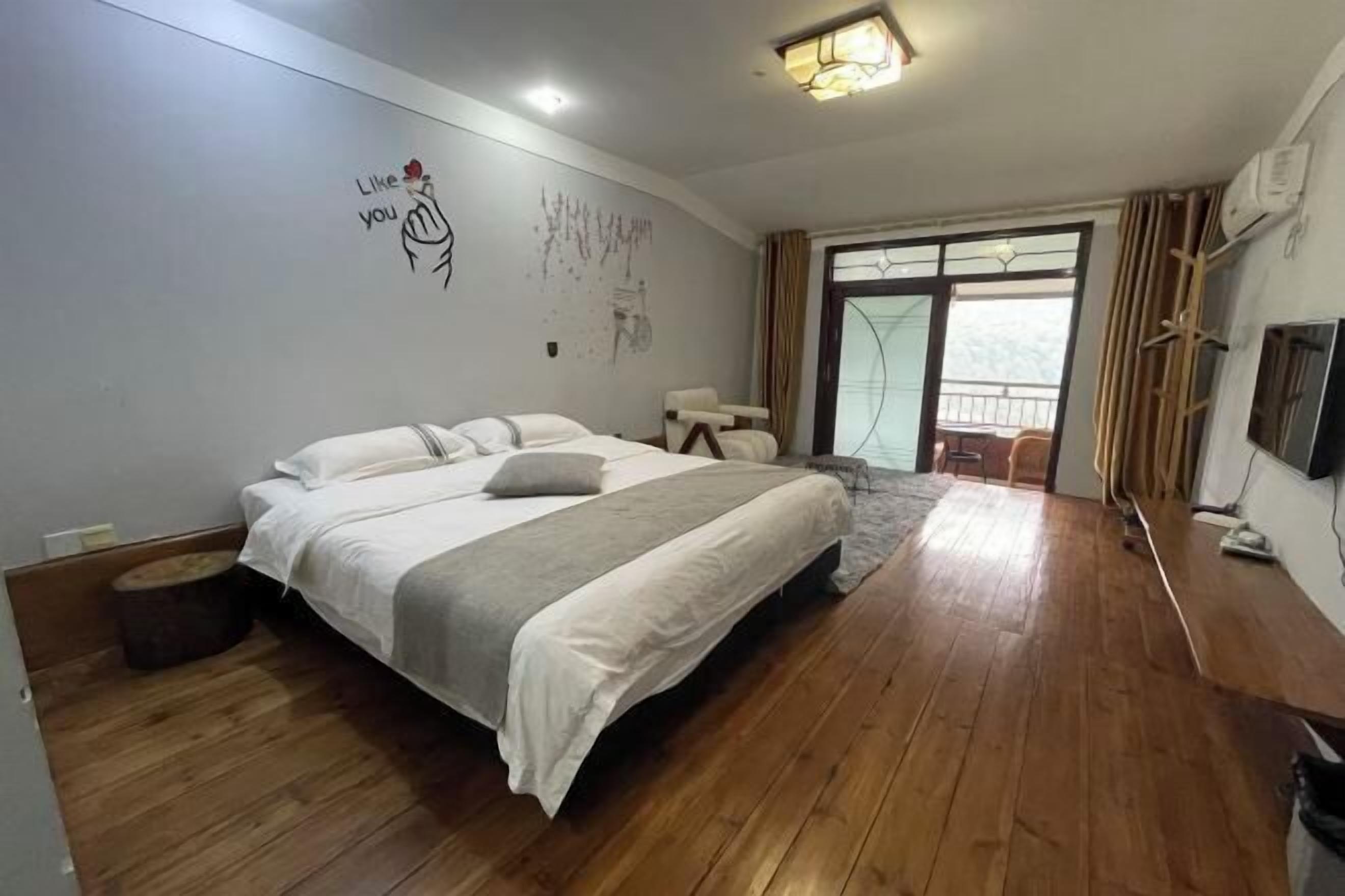 A bedroom with a large bed, wooden flooring, a window with curtains, and a wall with a drawing.