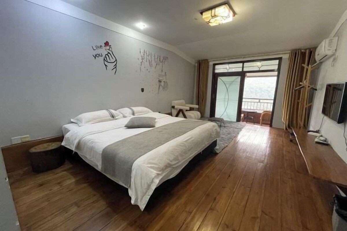 Een slaapkamer met een groot bed, houten vloeren, een raam met gordijnen en een muur met een tekening.